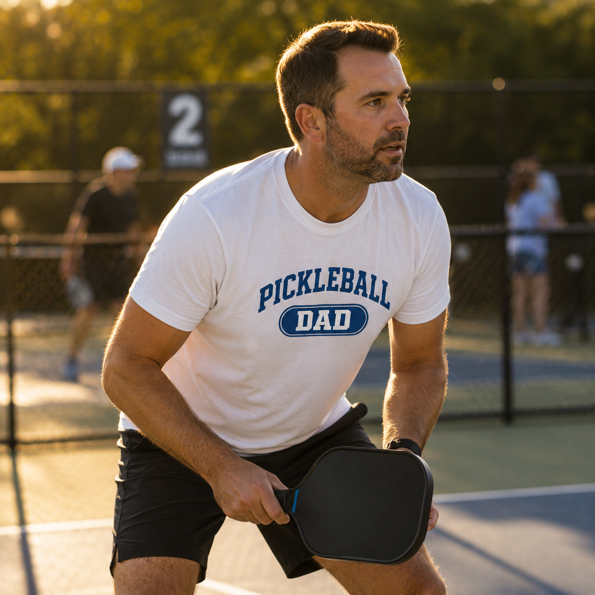 Pickleball dad wearing Pickleball Dad collegiate t-shirt playing on outdoor court in golden afternoon light