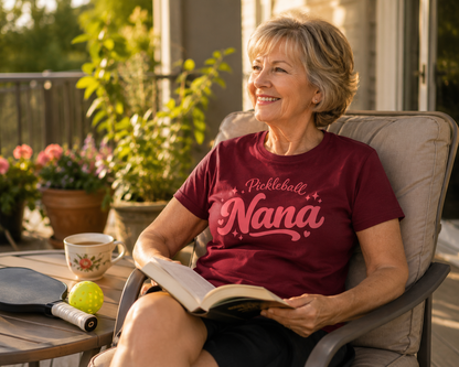 Woman wearing Pickleball Nana shirt reading on sunny terrace with paddle nearby