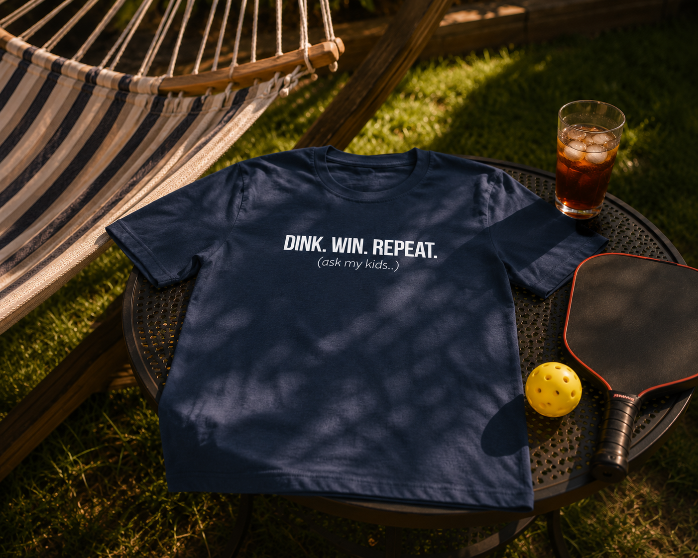 Dink Win Repeat Ask My Kids shirt folded on outdoor table next to hammock and paddle