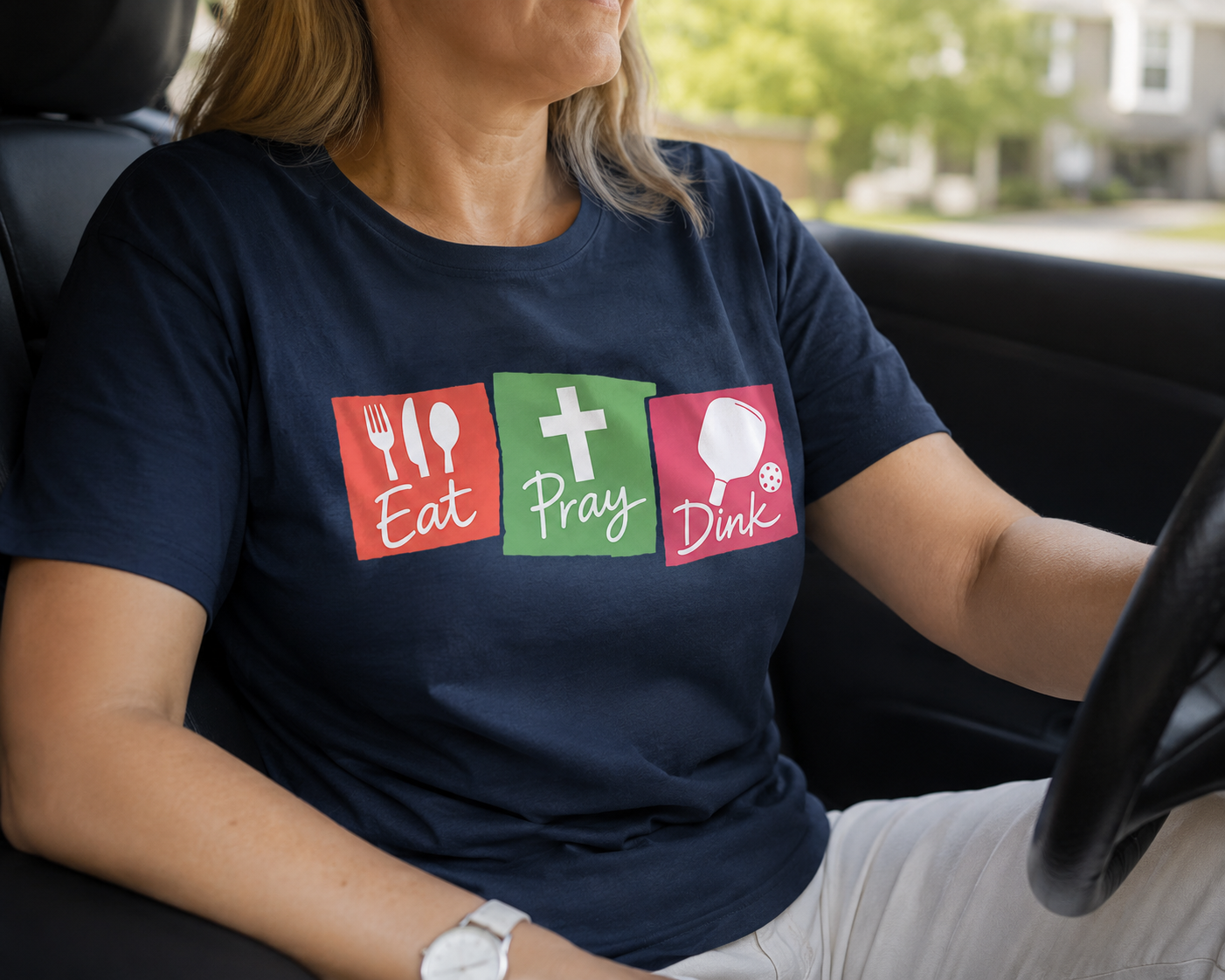 Woman in her 50s wearing Eat Pray Dink pickleball shirt driving a car